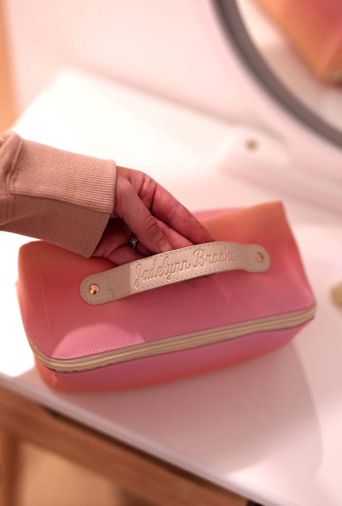 Pink cosmetic bag with cream handle, featuring embossed logo for stylish organization.