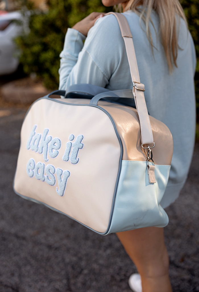 Stylish travel bag in cream, blue, and peach with embroidered 'take it easy' message and adjustable strap.