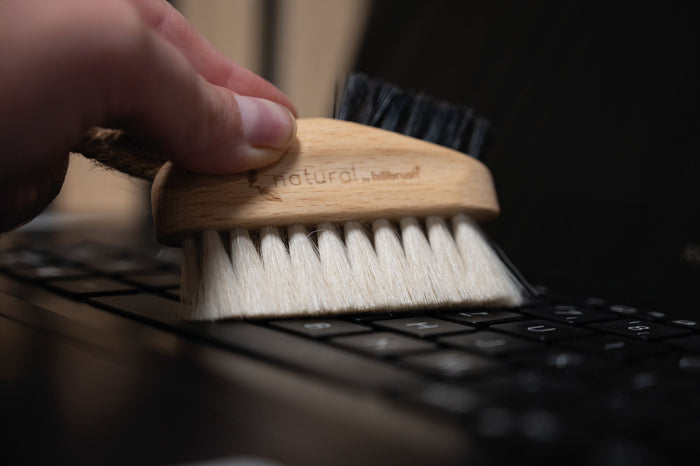 Hand holding a wooden brush with natural fibers over a keyboard