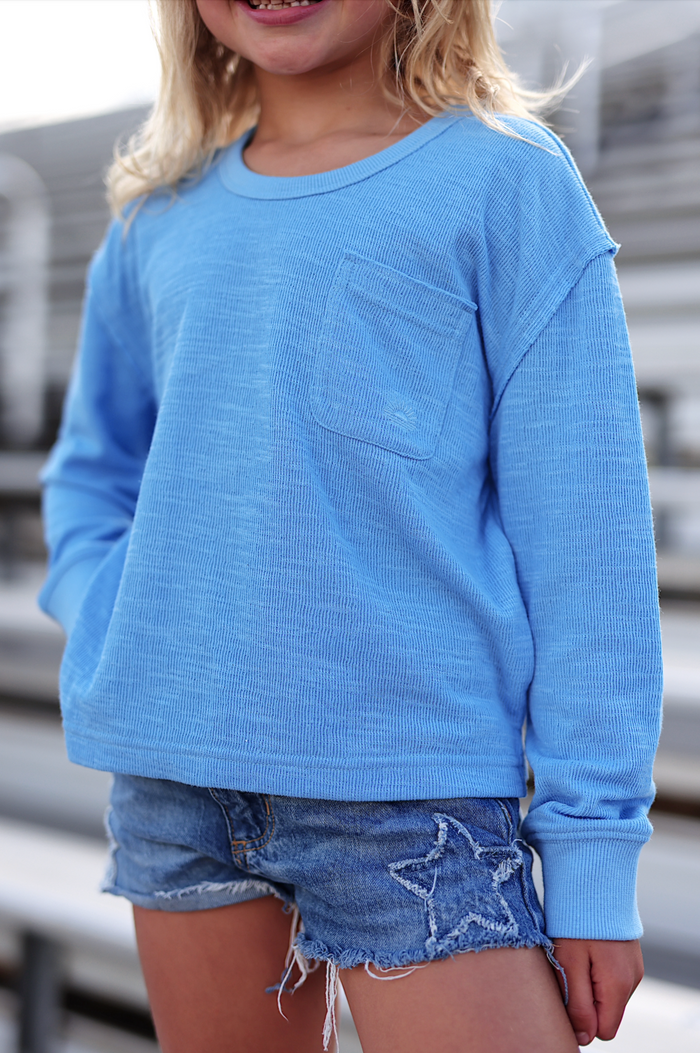 Light blue long-sleeve shirt for kids with pocket, paired with denim shorts for a stylish summer look.