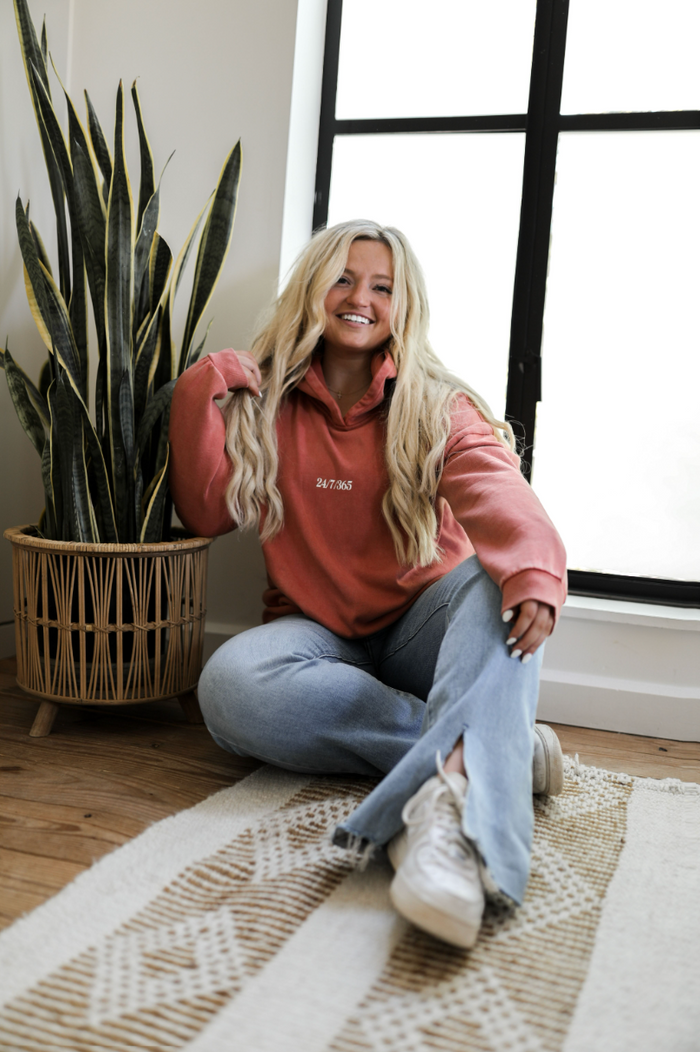 Women's coral hoodie with 24/7/365 print, paired with light blue flared jeans and white sneakers.