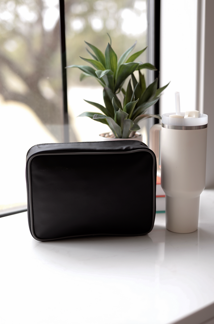 Black zippered cosmetic bag on a marble surface next to a white tumbler and green plant.