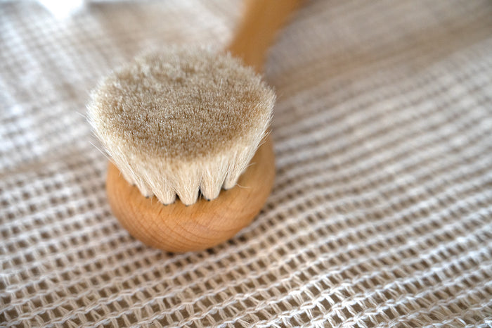 A close-up of a small wooden baby hair brush with soft beige bristles, resting on a woven beige fabric surface.