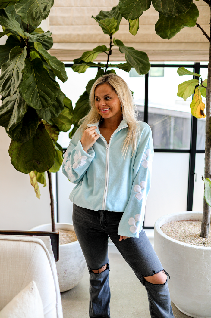 Light blue fleece zip-up jacket with floral designs on sleeves, paired with black ripped jeans.