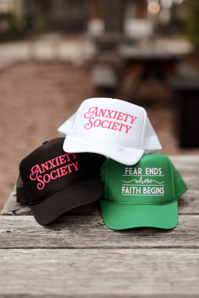Three trucker hats: black with 'Anxiety Society', white with 'Anxiety Society', green with 'Fear ends where faith begins'.