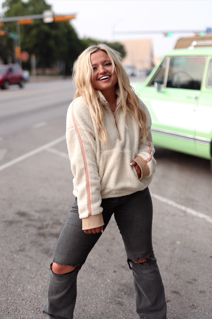 Cream fleece pullover with half-zip and pink stripes, worn with distressed black jeans.