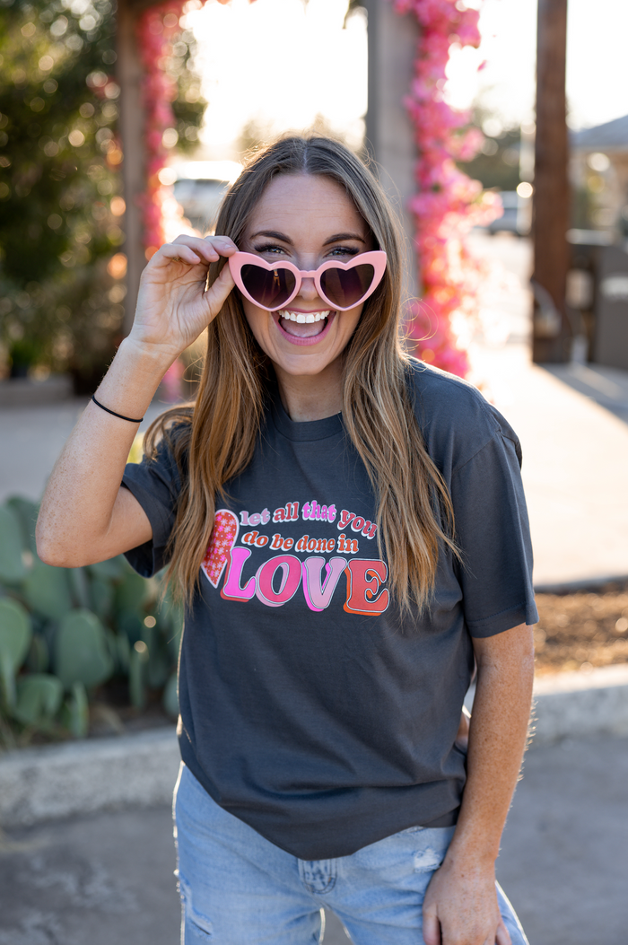 Woman wearing a gray graphic tee with 'Love' print and heart-shaped pink sunglasses.
