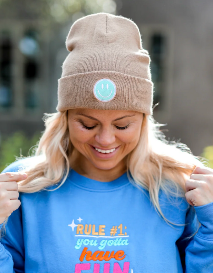 Smiley face tan beanie worn by a woman with blonde hair, paired with a blue sweatshirt.