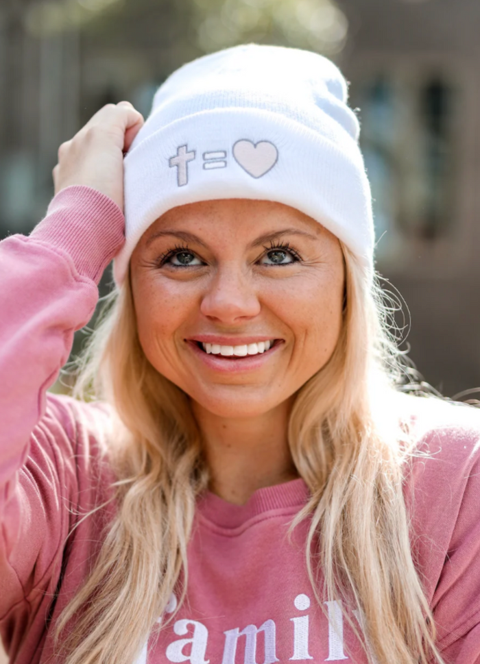 White beanie featuring cross and heart design, worn by smiling woman in casual pink sweatshirt.