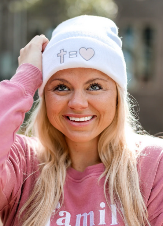 White beanie featuring cross and heart design, worn by smiling woman in casual pink sweatshirt.