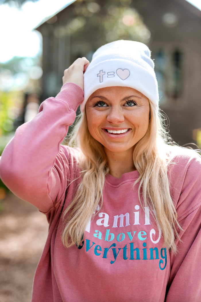 Woman wearing a white beanie with a heart and cross design, in a pink sweatshirt with 'Family above Everything' text.