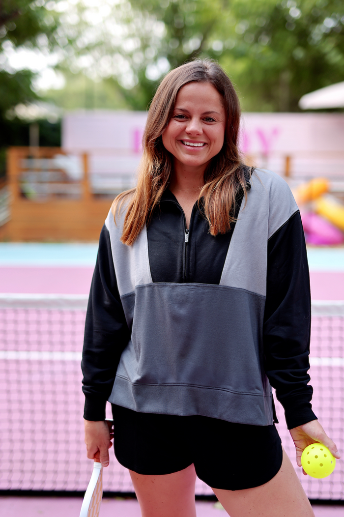 Woman smiling in a gray and black zip-up sweatshirt, holding a yellow pickleball, ready for sports.
