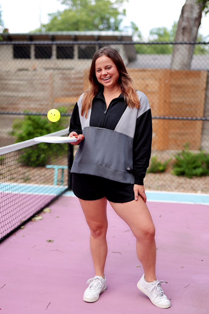 Woman in a black and gray athletic pullover with shorts, playing pickleball outdoors.
