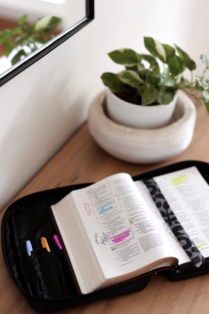 Open hardcover Bible with colorful highlighters in a black zip case on a wooden table.