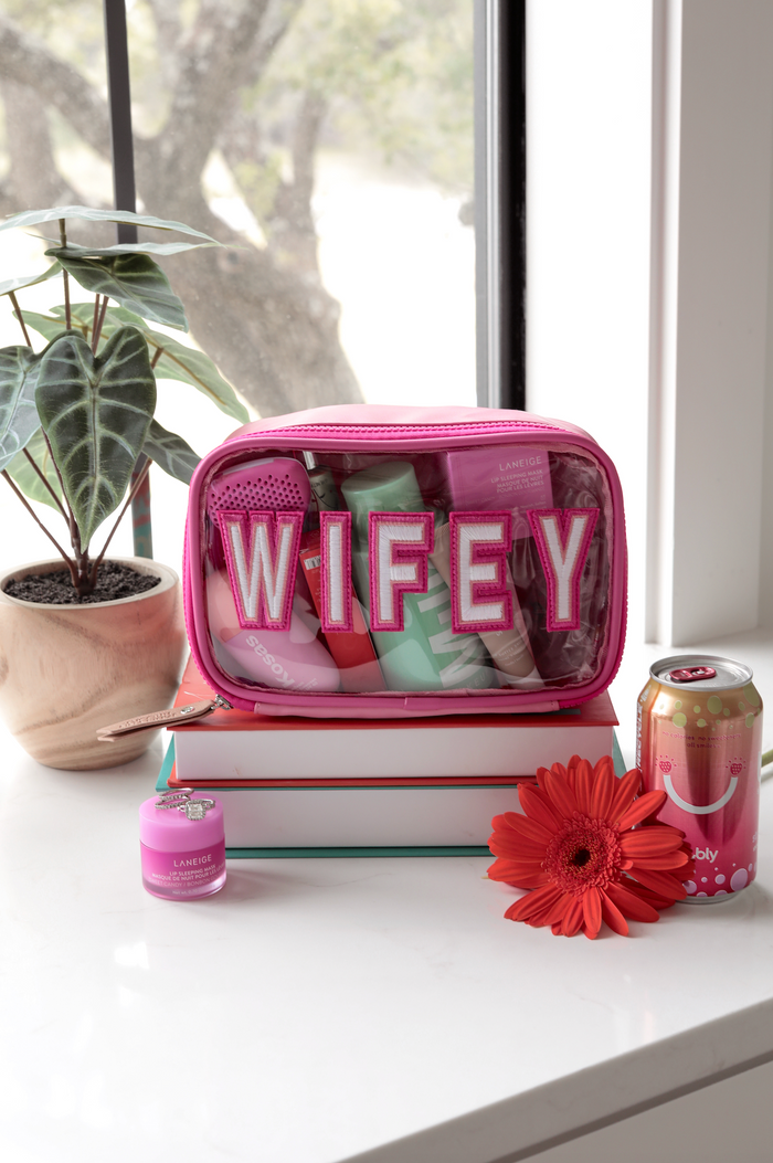 Pink cosmetic bag labeled 'WIFEY' filled with skincare products, flowers, and a sparkling drink.