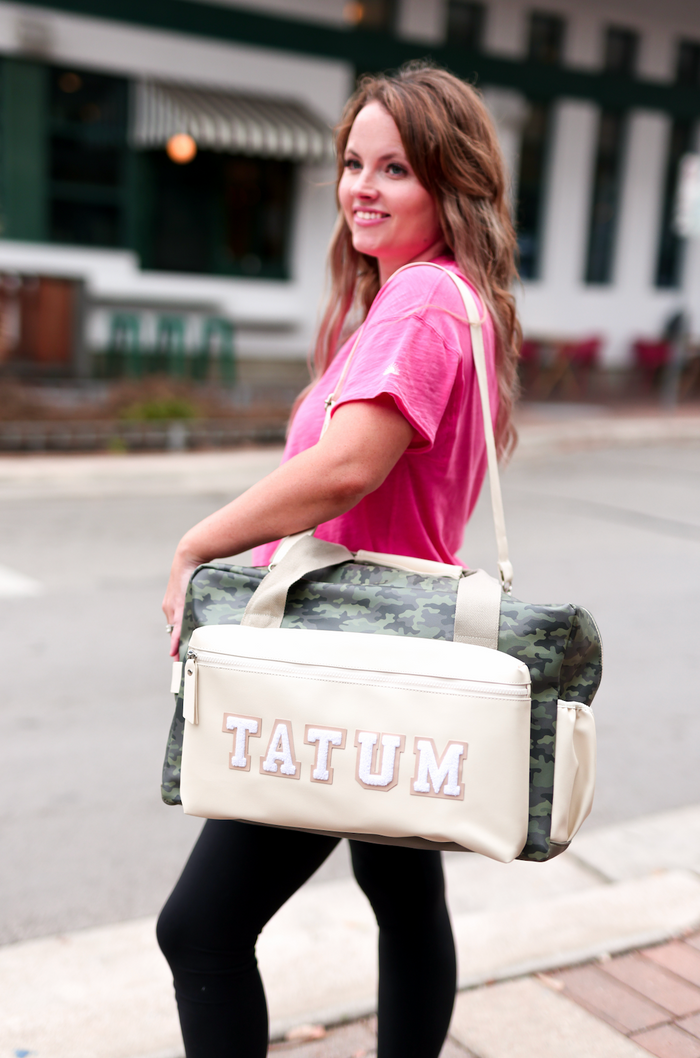 Woman holding a beige and camo duffel bag with 'Tatum' branding on a street.