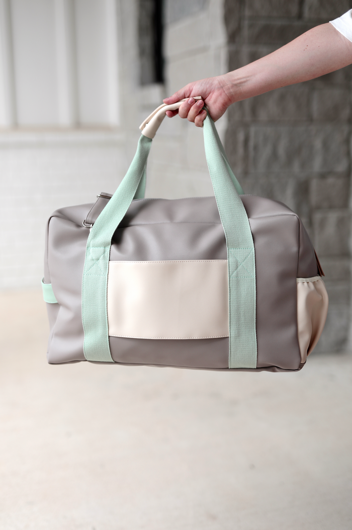 Stylish dual-tone duffel bag in gray and cream with mint green straps, made from durable material.