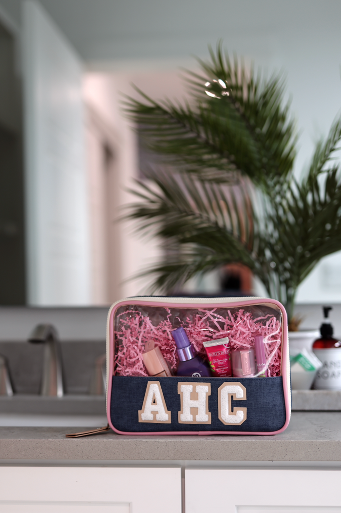 Stylish denim makeup bag with clear top, filled with beauty products in pink shredded paper.