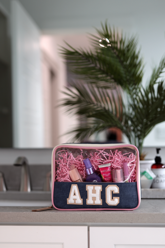 Stylish denim makeup bag with clear top, filled with beauty products in pink shredded paper.
