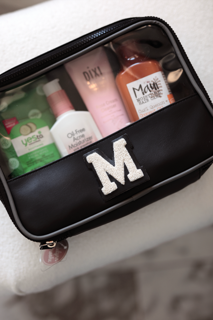 Black zippered cosmetic bag with clear top and personalized 'M', holding skincare products.