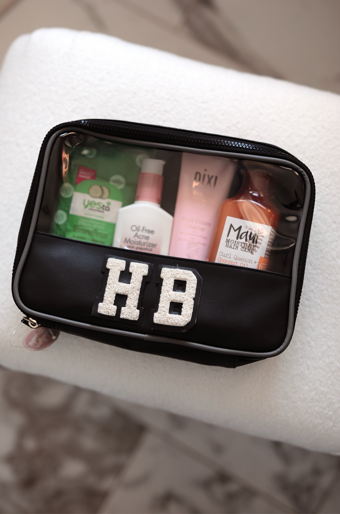 Black cosmetics bag with clear top, includes skincare and hair products, personalized with 'HB' embellishments.