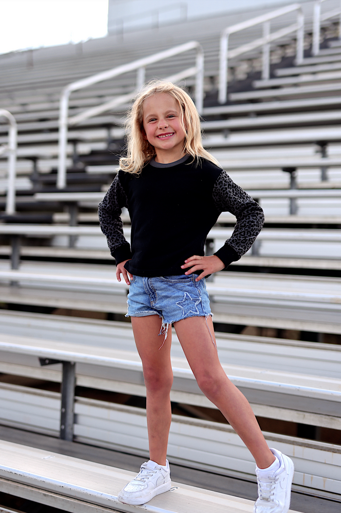 Girl wearing black leopard-sleeve sweatshirt and denim shorts, perfect for casual outings.