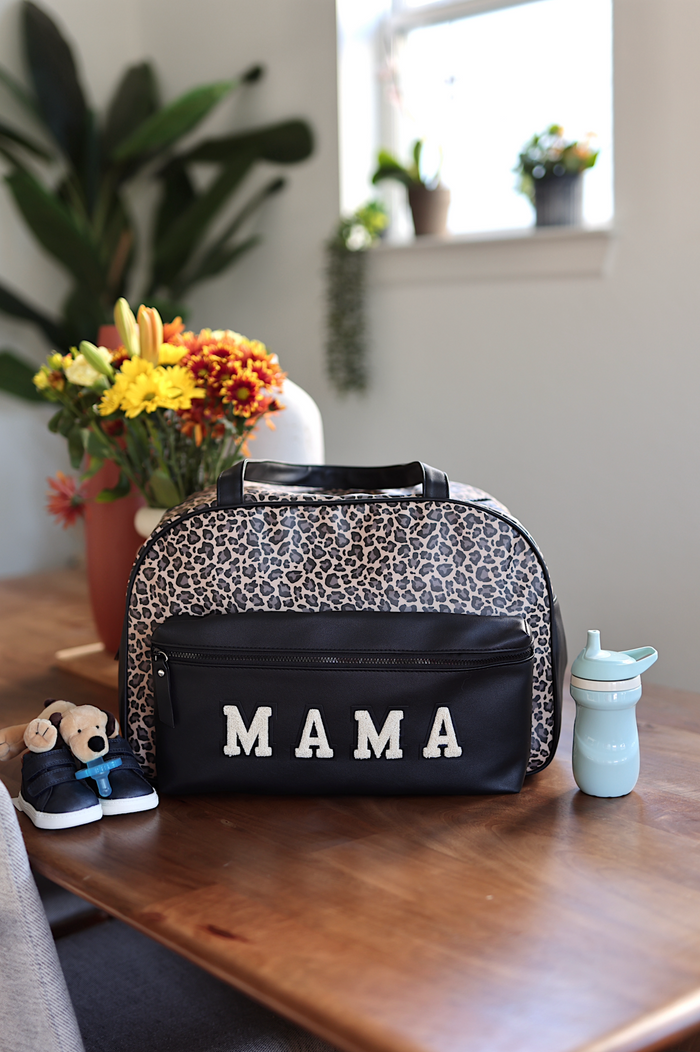 Leopard print bag with 'MAMA' on a wooden table next to a teddy bear and sippy cup.