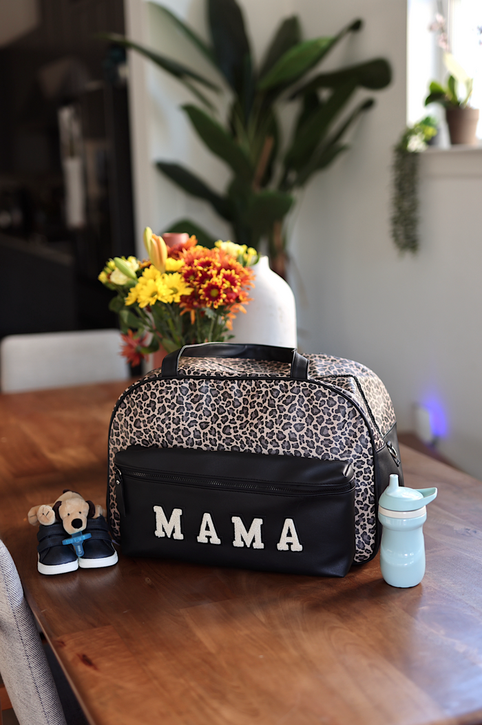Leopard print bag with 'MAMA' on a wooden table with a teddy bear and water bottle.