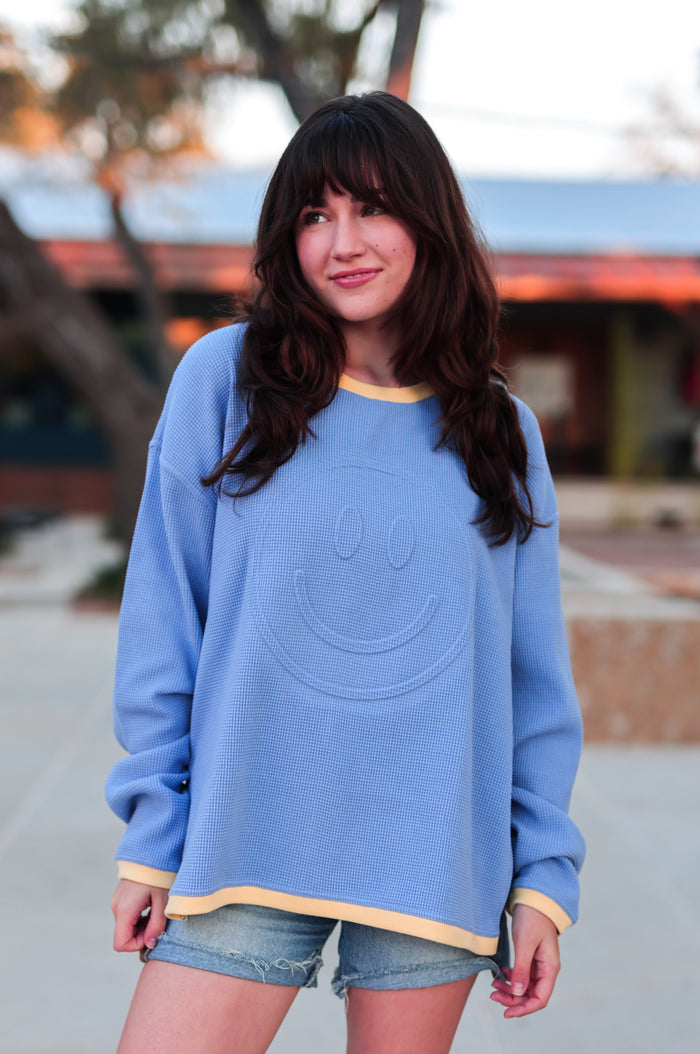 Woman wearing a blue and yellow waffle sweatshirt with a embossed smiley with trees around.