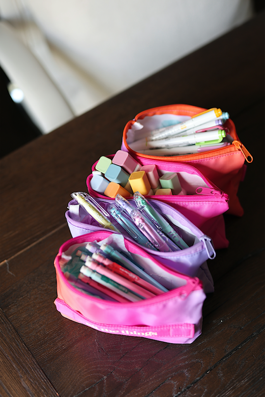 Colorful zippered pencil cases in orange, pink, and purple filled with pens and supplies.