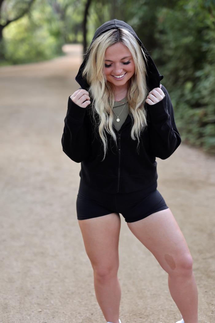 Woman wearing a black zip-up hoodie and black shorts in a natural outdoor setting.
