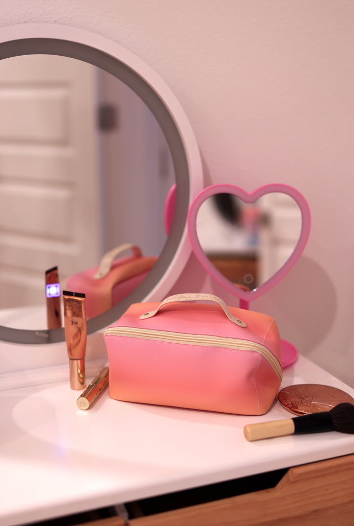 Pink gradient makeup bag with zipper, surrounded by beauty products on a vanity.