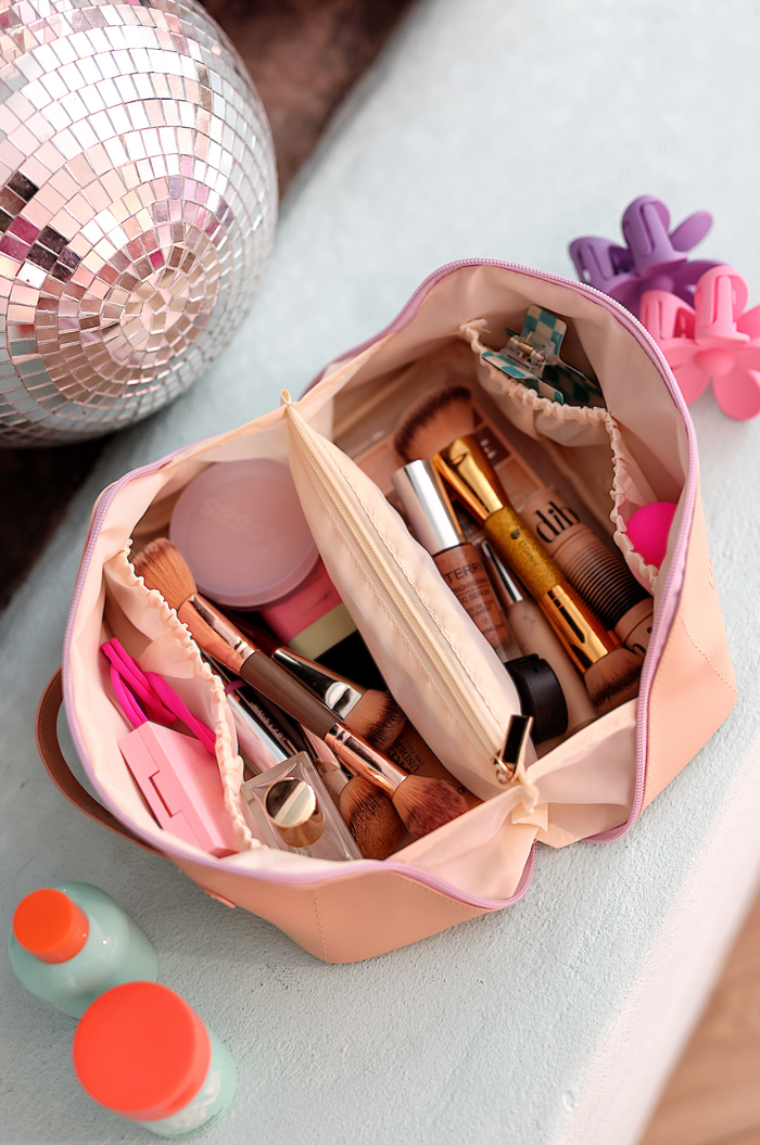 Light pink makeup bag filled with cosmetics and brushes, featuring multiple compartments for organization.
