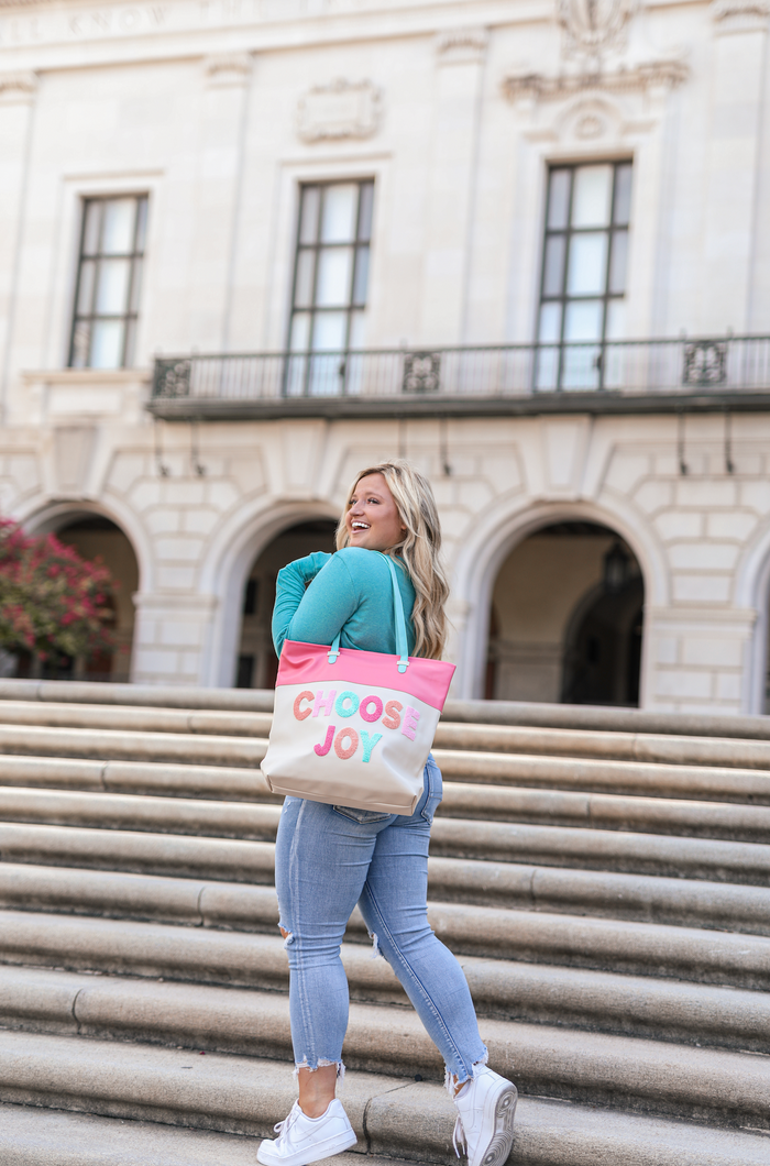 Woman in blue sweater carrying a 'Choose Joy' tote bag on stairs, showcasing pink and cream colors.
