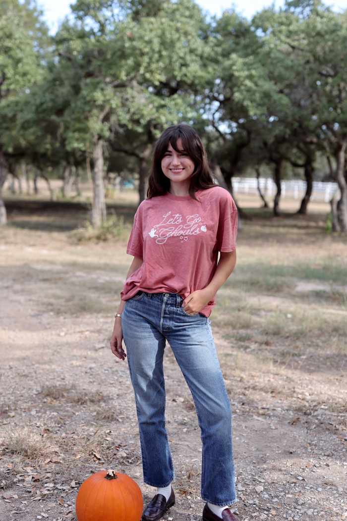 Woman wearing a pink t-shirt and blue jeans standing in a field with trees in the background