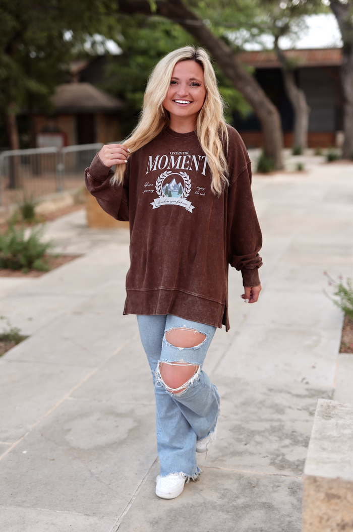 Brown oversized sweatshirt with 'Live in the Moment' graphic, paired with distressed light blue jeans.