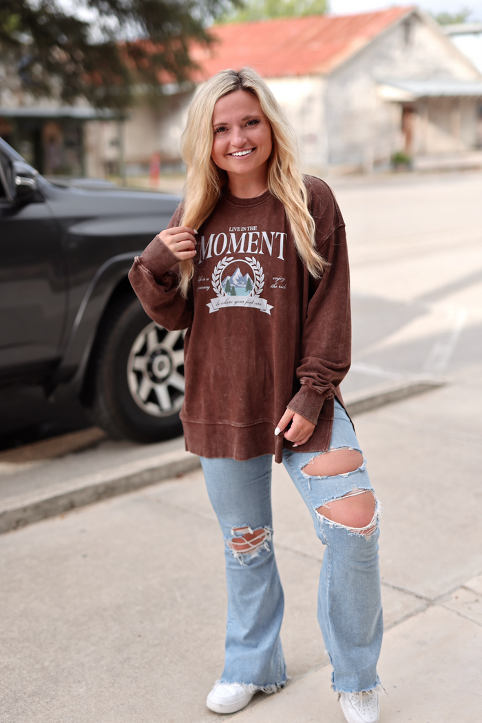 Brown graphic sweatshirt with 'Live in the Moment' design, paired with distressed flared jeans and white sneakers.