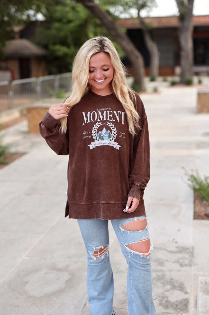 Brown oversized sweatshirt with 'Live in the Moment' graphic, worn with distressed blue jeans.