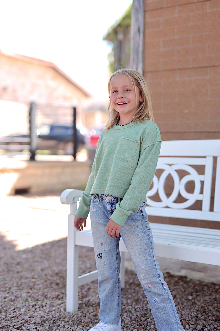 Child wearing a light green long sleeve shirt and stylish embroidered jeans, smiling outdoors.