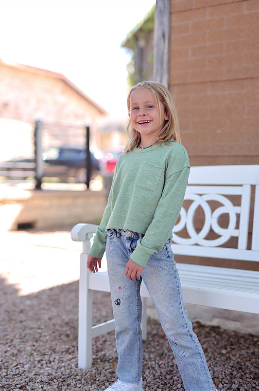 Child wearing a light green long sleeve shirt and stylish embroidered jeans, smiling outdoors.