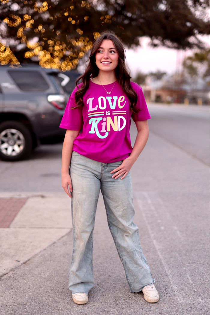 Pink graphic tee featuring 'Love is Kind' slogan, paired with light wash flared jeans and casual sneakers.