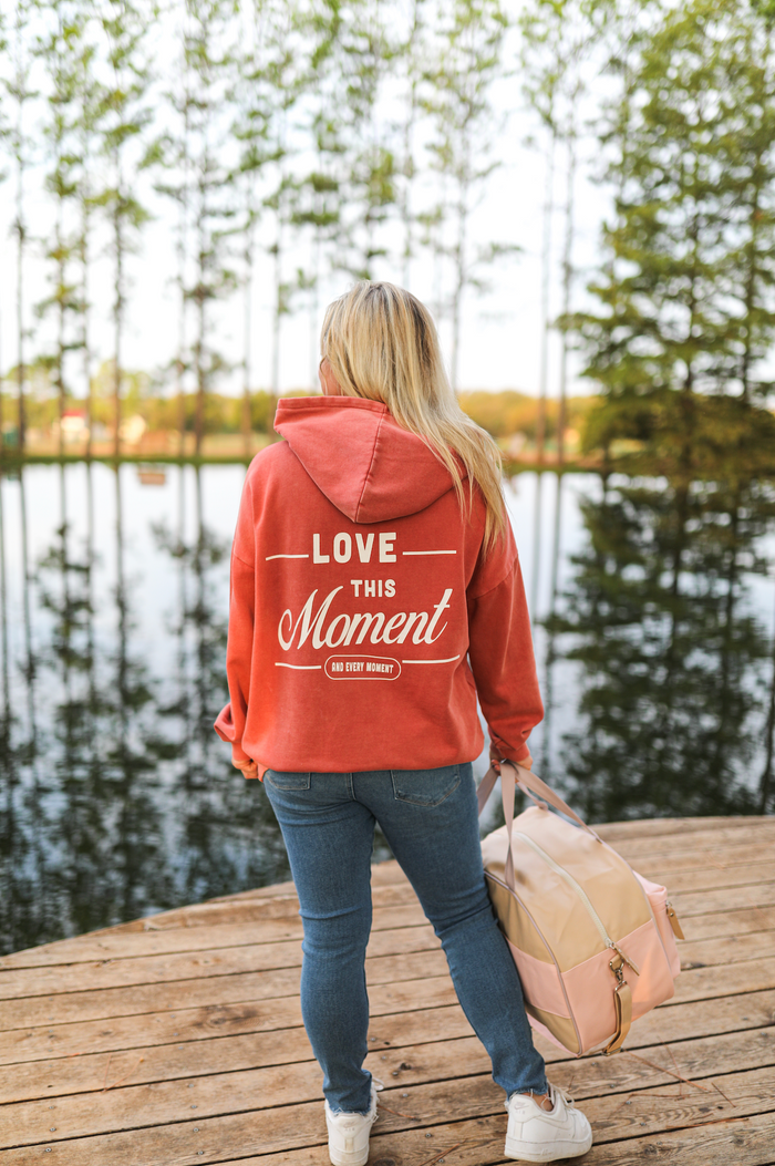 Women's coral hoodie with 'Love This Moment' print, paired with jeans by a scenic lake.