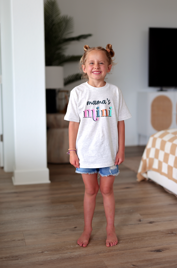 Girl wearing gray 'mama's mini' graphic t-shirt with denim shorts, standing indoors.