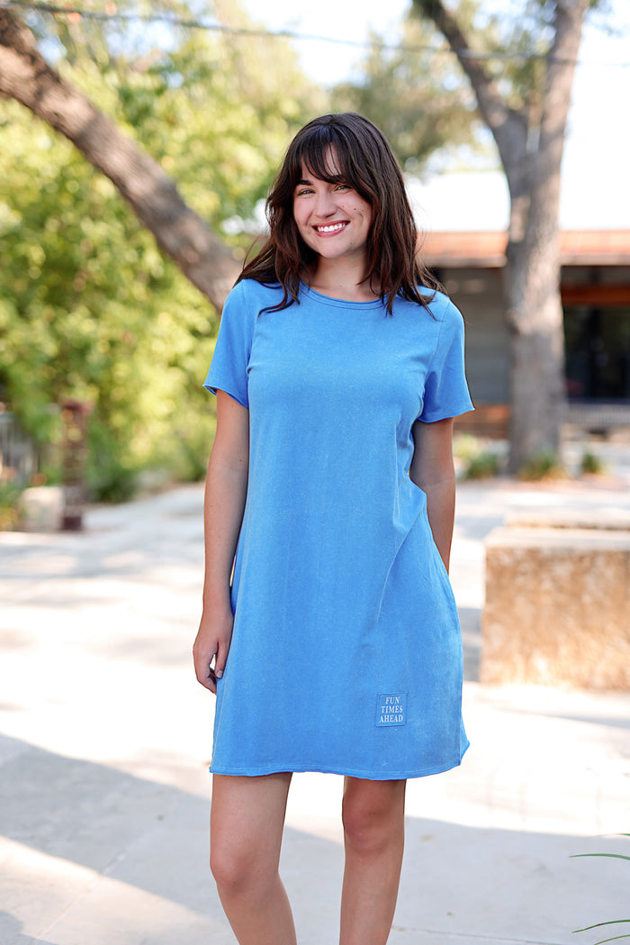 Woman wearing a blue dress standing outdoors with trees and a building in the background
