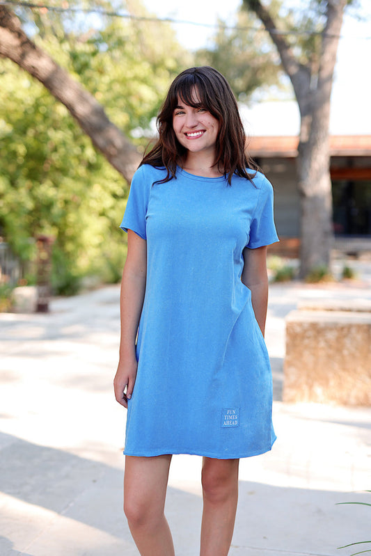 Woman wearing a blue dress standing outdoors with trees and a building in the background