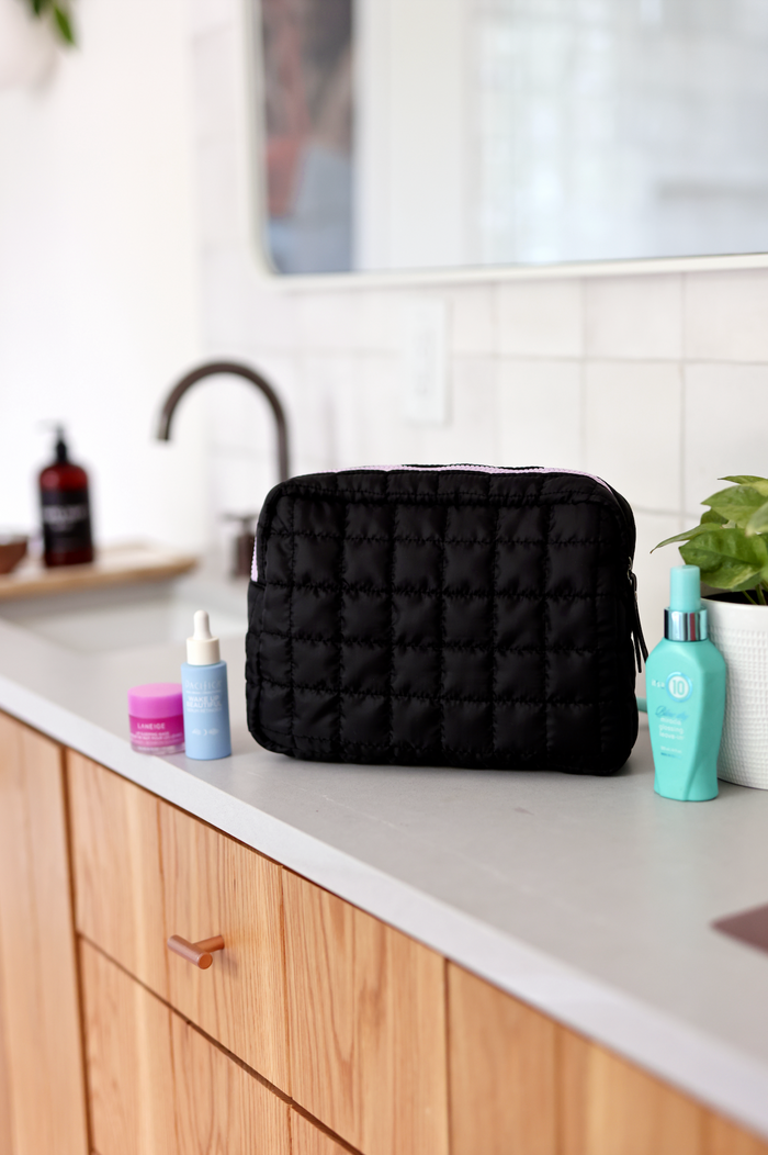 Black quilted cosmetic bag on a bathroom counter, paired with skincare products.
