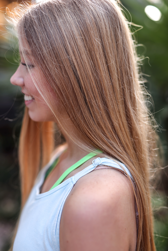 Side view of a woman with long blonde hair wearing a light blue tank top with green straps.