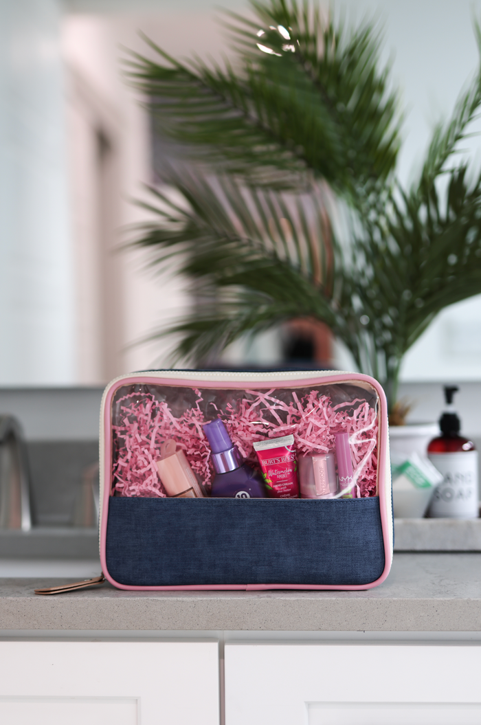 Clear cosmetic bag with denim base, pink trim, filled with beauty products and shredded paper.