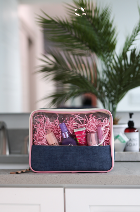 Clear cosmetic bag with denim base, pink trim, filled with beauty products and shredded paper.