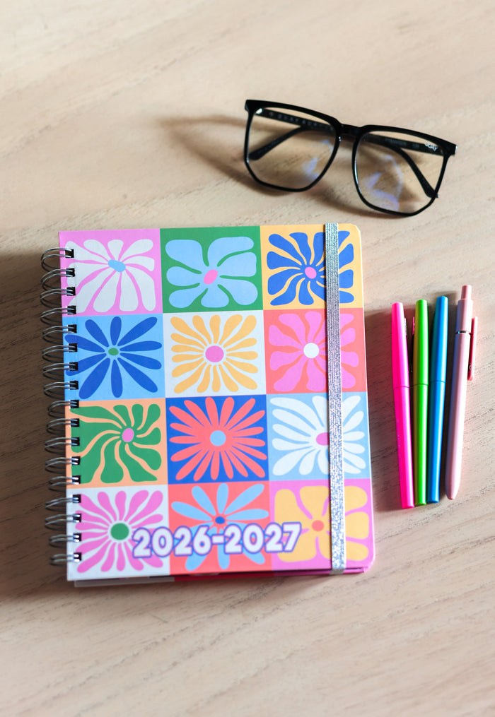Colorful planner with floral pattern and 2026-2027 text on a wooden surface with glasses and pens.